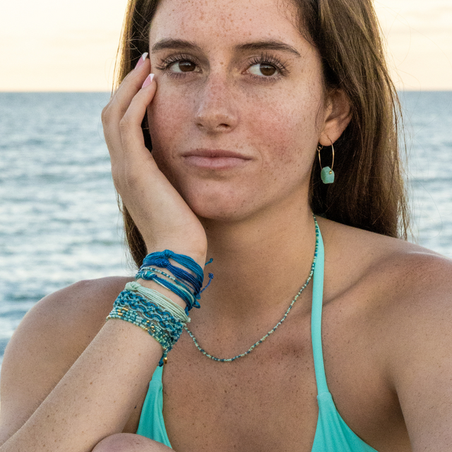 Model sitting on the beach wearing blue and teal bracelets, necklac, and earrings.