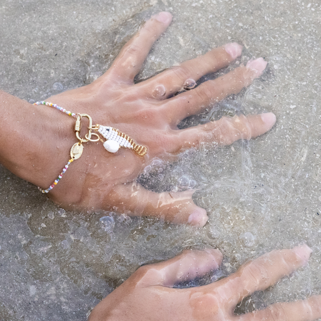 Hands in sandy waves wearing a charm bracelet with a white & gold fish charm and a pearl charm on a beaded bracelet with carabiner.