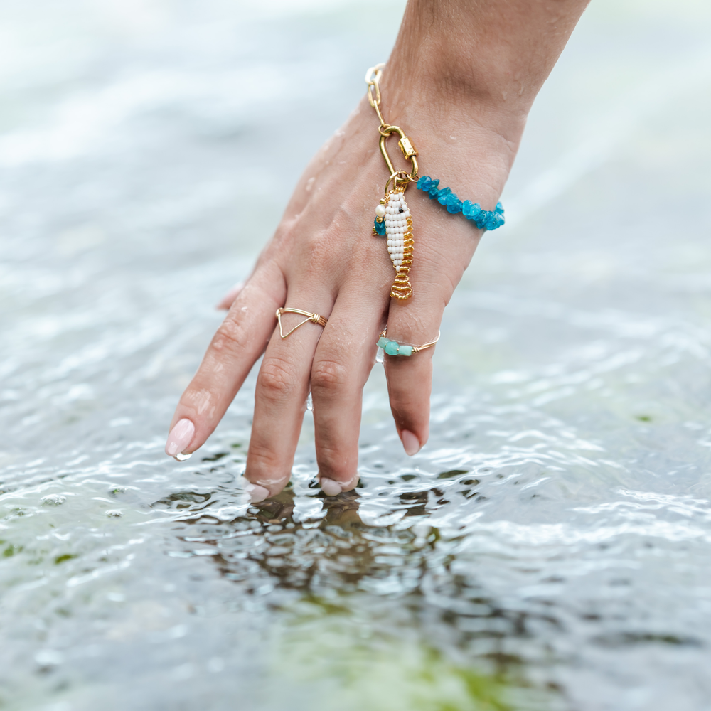 Hand wearing apatite charm bracelet with a white & gold beaded fish charm and stone charm worn with stone ring and triangle ring