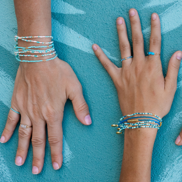 Two model's hands with teal stretch bracelets and rings with a painted teal leaf background