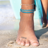 Surfer woman wearing string anklets in blue colors with one gofl filled bead stanging on the sand with teal surfboard