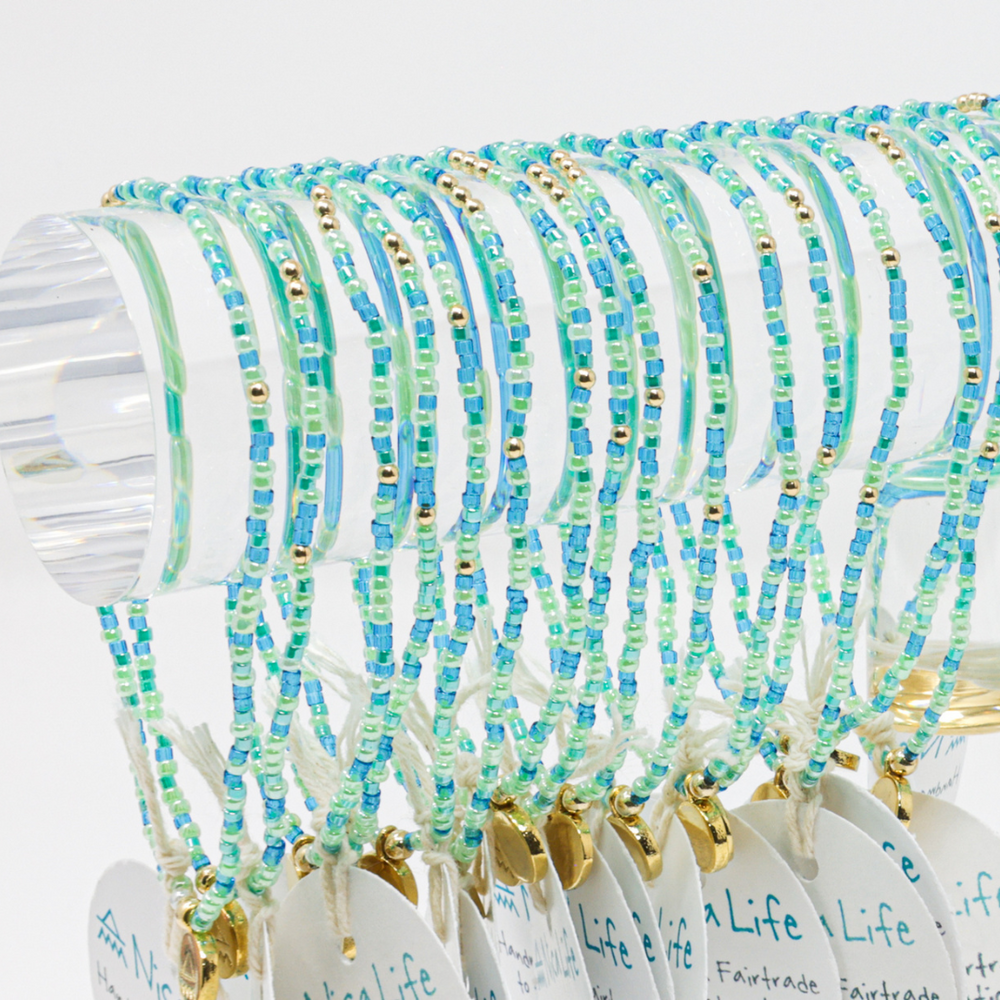 Many blue green and teal beaded bracelts with gold beads shown on an acrylic bar with a white background.