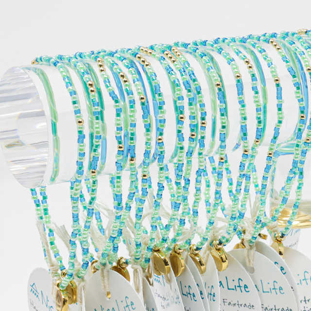 Many blue green and teal beaded bracelts with gold beads shown on an acrylic bar with a white background.