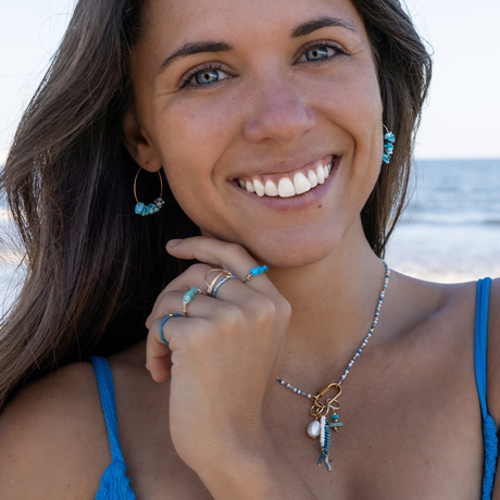 Woman wearing charm bar necklace with fish charms, rings & earrings all in blue colors on the beach