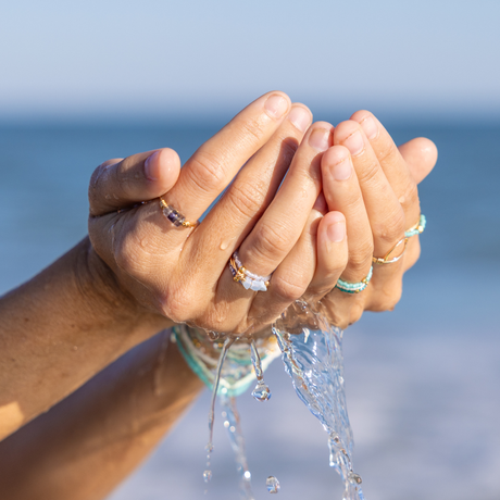 Close-up of hands with purple& teal stacking rings holding water with a blurred ocean background