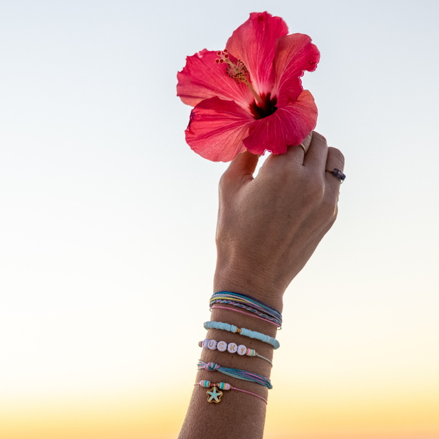Colorful bracelet stack with bead and string and charms shown on a woman holding a pink/red flower in the air