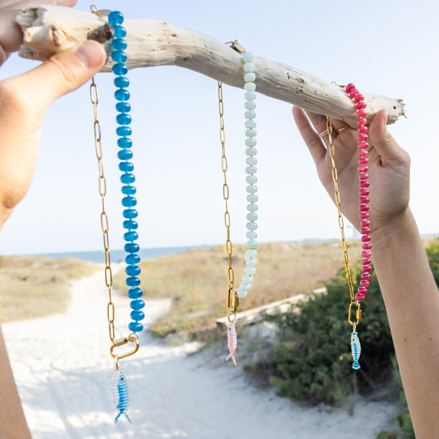 Three charm bar necklaces made with colorful stones with colorful fish charms shown on a piece of driftwood with beach background