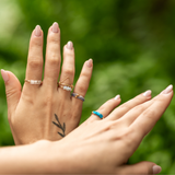 Model holding her hands up with green tropical background wearing stacking rings of different kinds. Stone rings, a triangle ring, and stretch rings. 