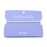 White & gold Nica Life bag charm on a purple card with brand name.