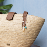 Woven straw bag with brown leather handles and a white & gold bag charm on a light blue background