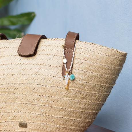 Woven straw bag with brown leather handles and a white & gold bag charm on a light blue background